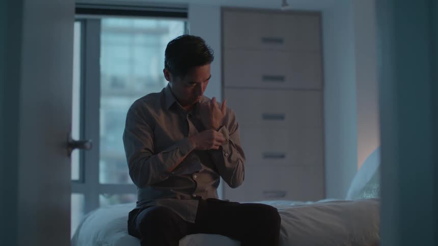 A man sits on his bed and buttons his shirt up as he gets dressed