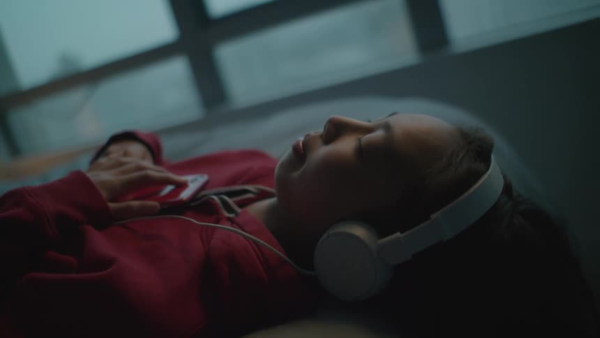 A girl in a room with headphones listening to a smart phone with a wall and window in the background
