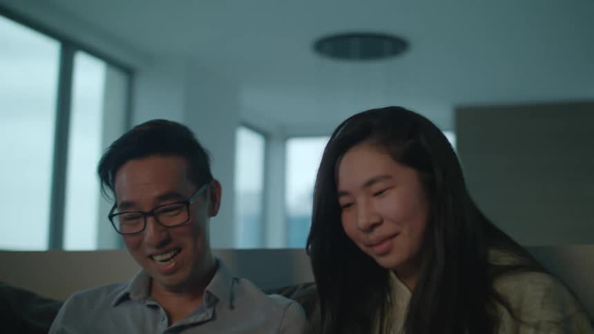 A man and a woman in a room on a couch looking at a laptop with a wall and window in the background
