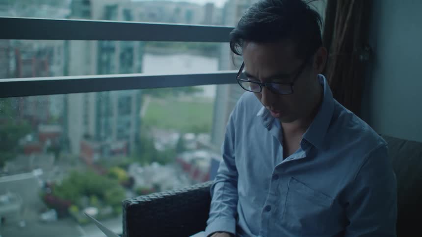 An asian male is sitting in a chair while on a laptop talking and typing