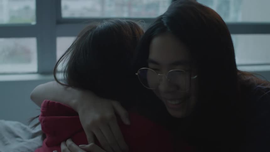 Two women smile and hug while one is sitting on a bed
