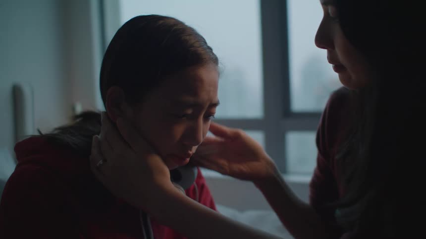 A woman in a room hugging and kissing a boy with headphones with a wall and window in the background