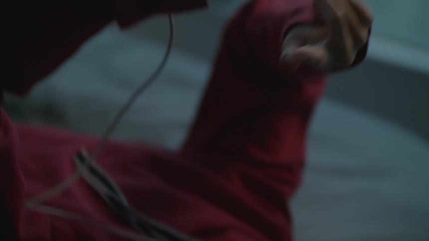 A young woman listening to music on a smartphone through her headphones uses a finger to scroll the screen as she lays on a bed