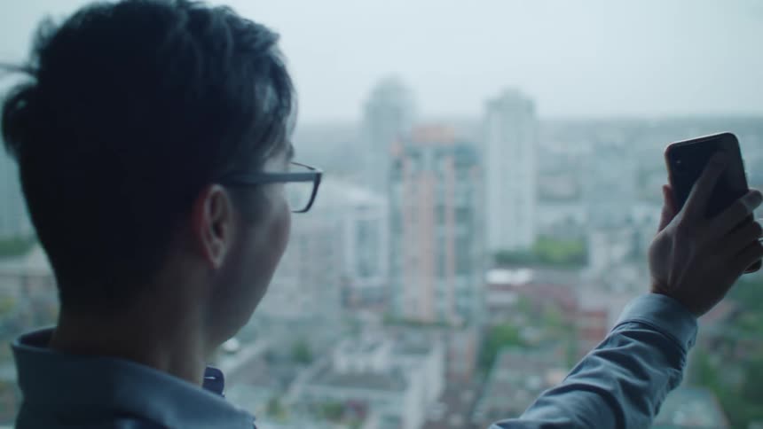 Man with glasses and blue shirt laughs and makes video call on smartphone showing the city buildings with front camera