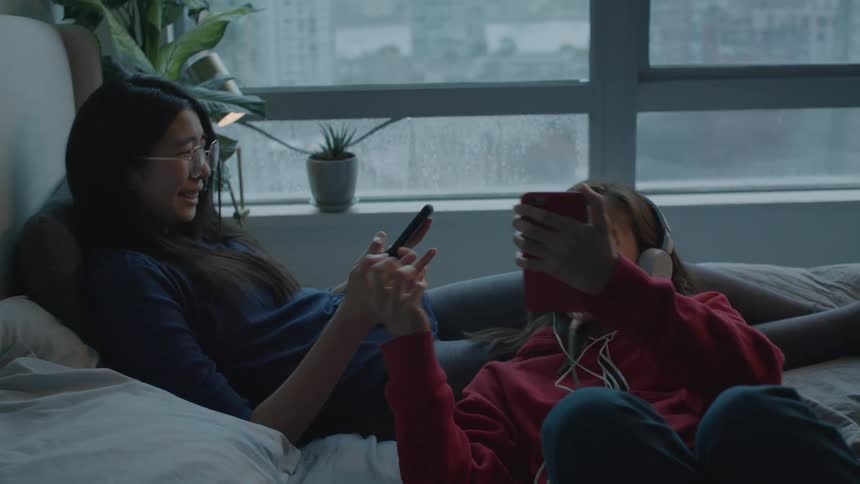 Women lie on bed playing with tablet and smartphone before showing each other their screens with a smile