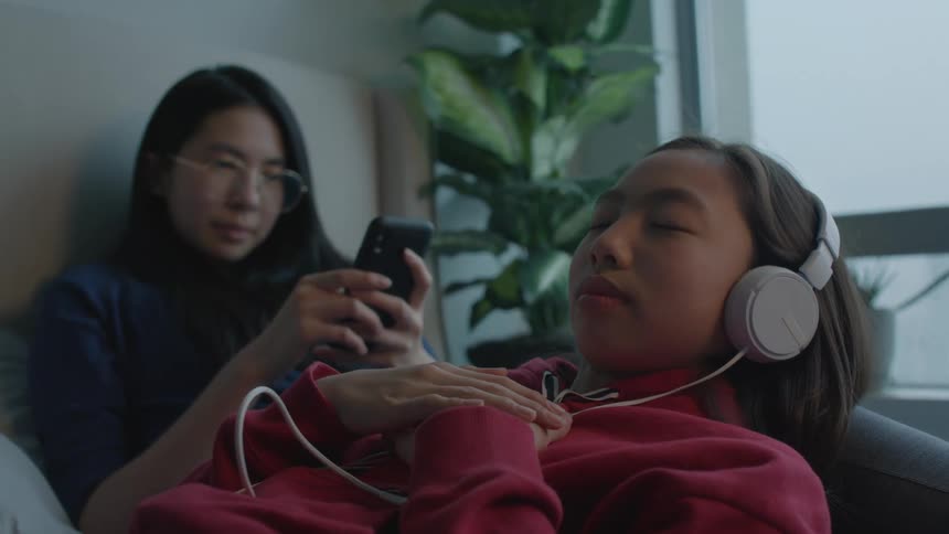 A girl leans on her sisters leg and listens to music and her sister who is using her cell phone bumps her leg up and laughs