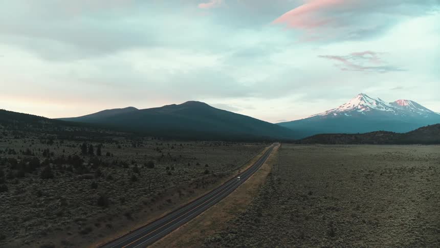 Lone vehicles rides along long road through scrub land away from mountains under red tinted sky