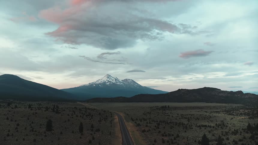Road passes through conifer dotted plains and hills toward twin peaked snow covered mountain under red tinted clouds