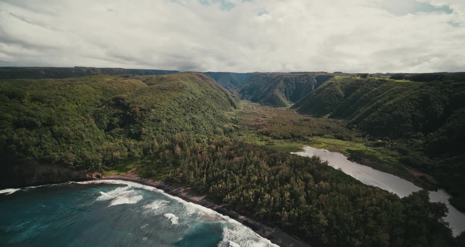 Aerial view of a coastline as waves roll in near a trees and mountains under a cloudy sky