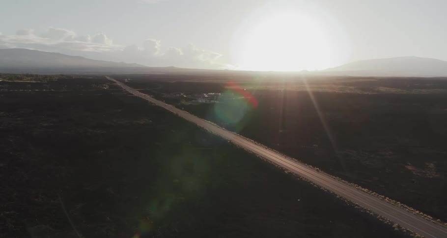 Sun glares from horizon through mist as highway passes through fields past buildings