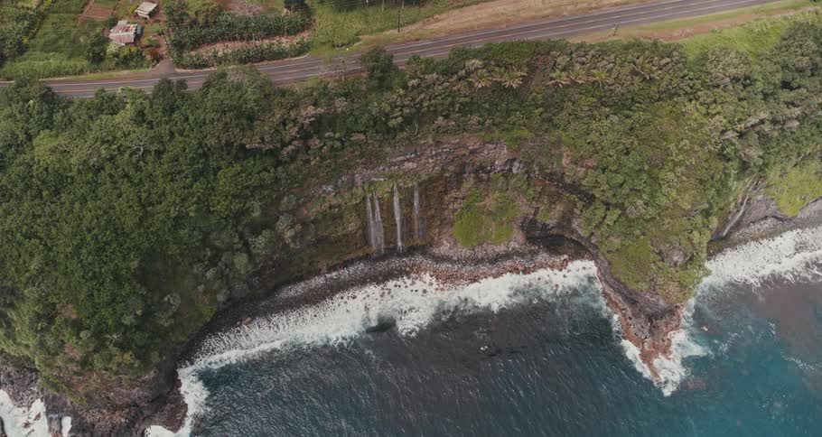 Aerial view of a coastline with waves rolling in and waterfalls flowing down a cliff lined with trees