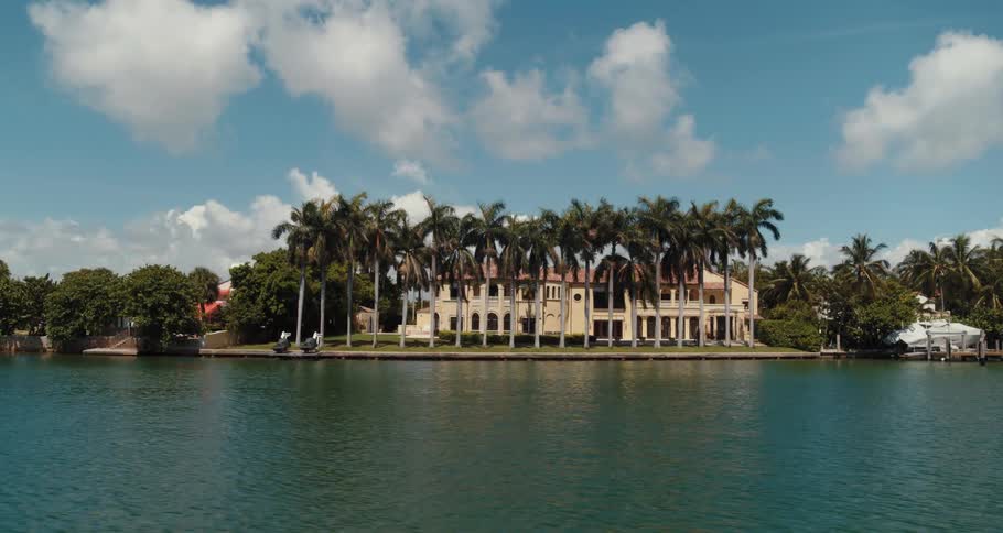 River water reflects palm trees and large home under cloudy sky