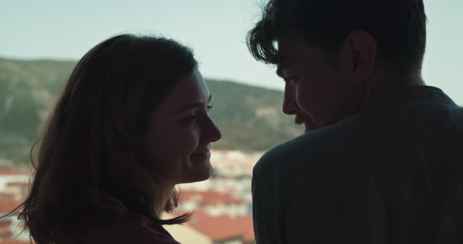 Couple smile at each other and kiss with red roofed town and green hill in background under a clear blue sky