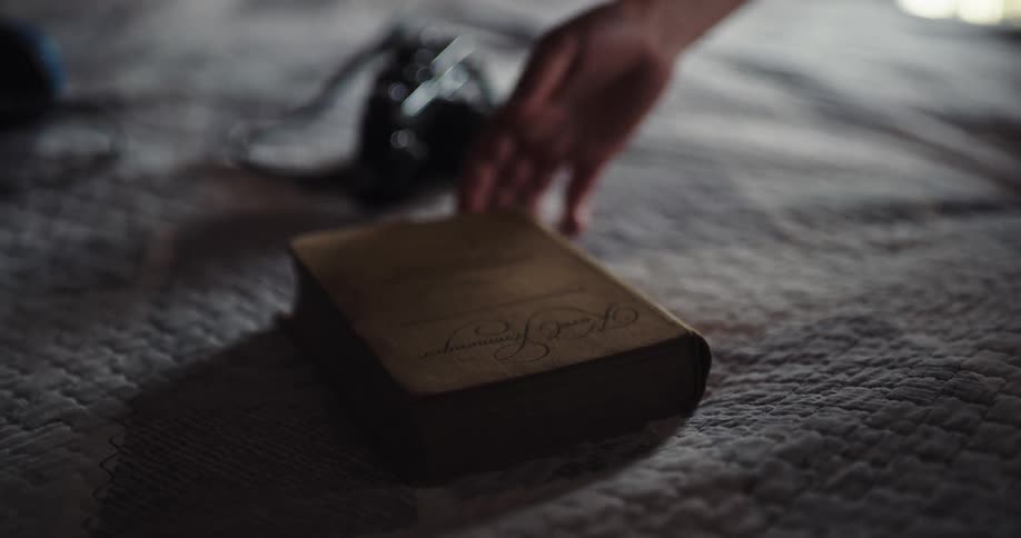 A person picking up sunglasses and a book from a bed