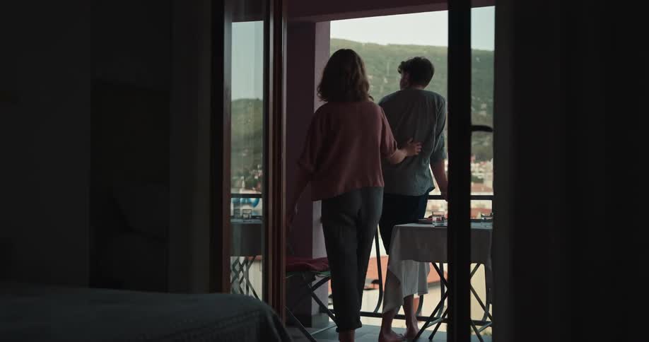 Woman joins man standing at hotel room balcony overlooking red roofed town and low mountain before kissing him