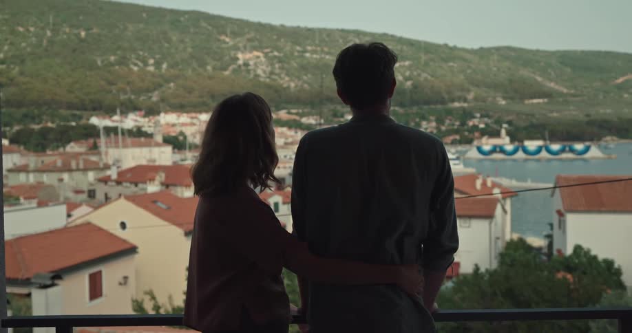 A woman joins a man on a balcony overlooking a bay and kisses him