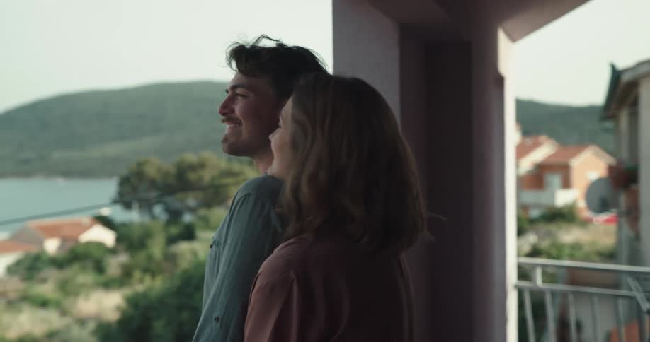 Woman with brown hair cuddles man with goatee beard watching coastal town from balcony