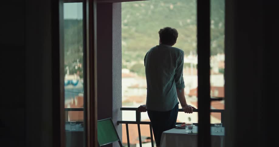 Man with shirt and goatee beard looks around from the balcony in the coastal town