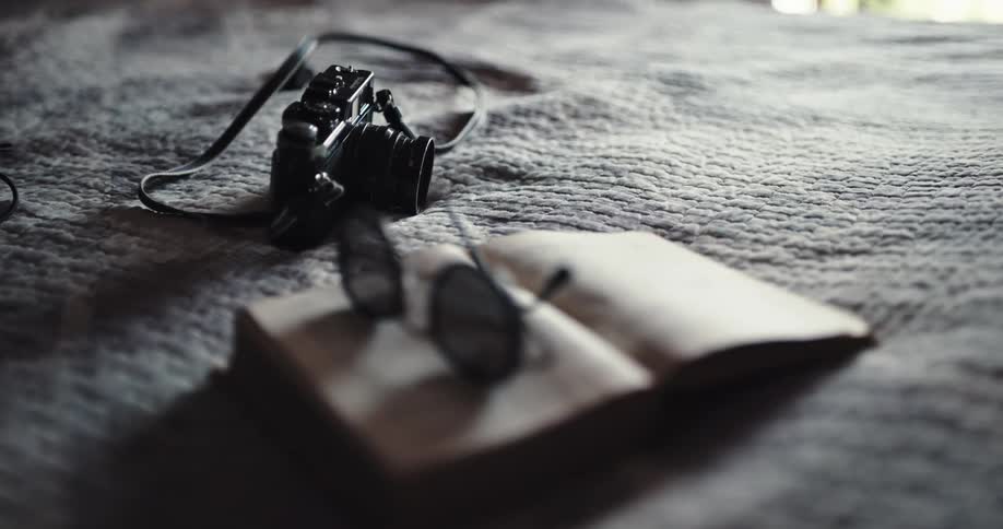 A pair of glasses sitting on an open book and a camera on a bed