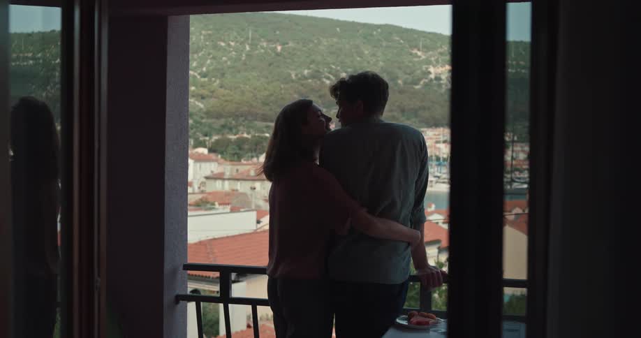 A woman wraps her arm around her boyfriend as he stands on a balcony and leans in for a kiss
