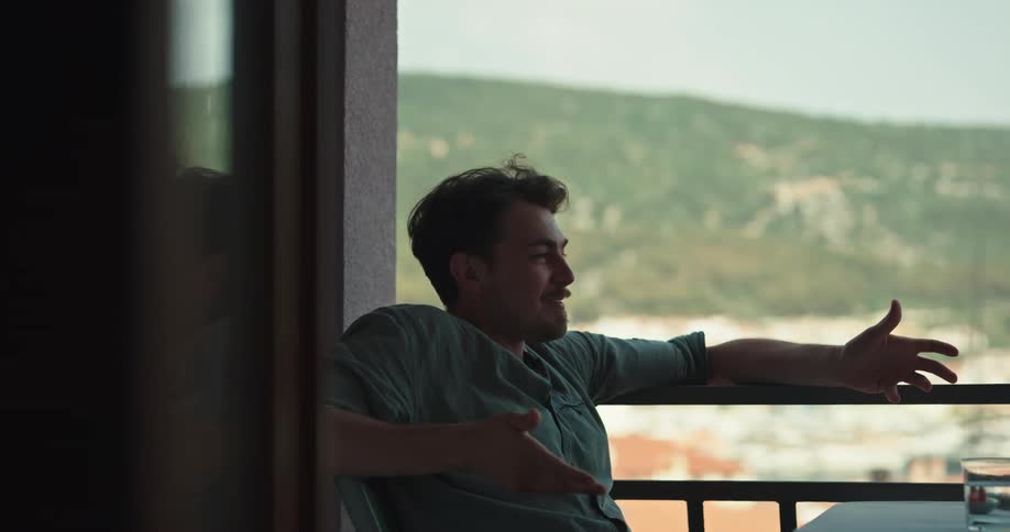 Man with goatee beard and blue shirt leans on balcony balustrade and talks to person