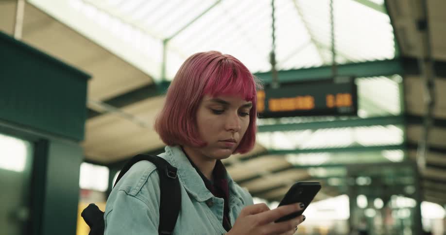 Woman with hot pink dyed hair plays on mobile phone while waiting at transit terminal as yellow train rolls through station