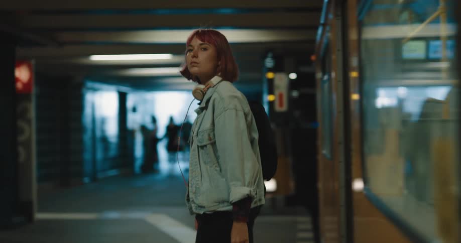 A woman with headphones and a backpack walking out of a train onto a train station
