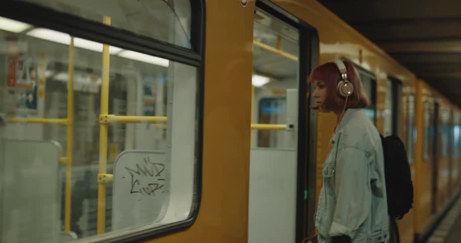 Woman with dyed hair wearing headphones and tall man wearing face mask get on and off train car respectively as train sits at terminal