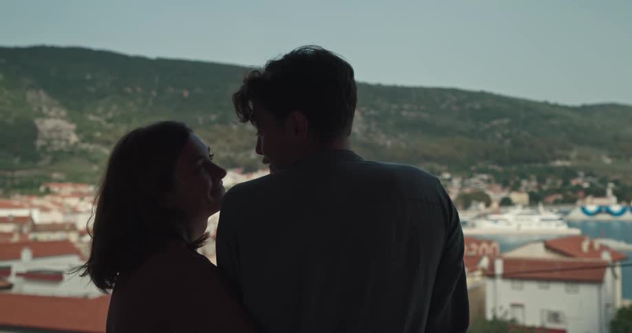 Couple hug and kiss on balcony overlooking red roofed town nestled between low mountain and water