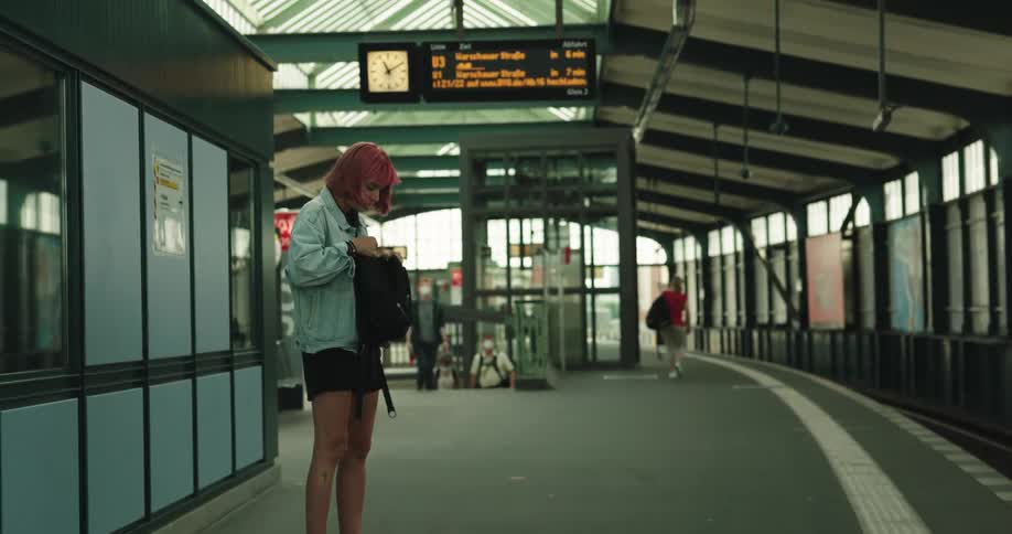 A woman searches her bag and pulls out a book while standing on a train station platform
