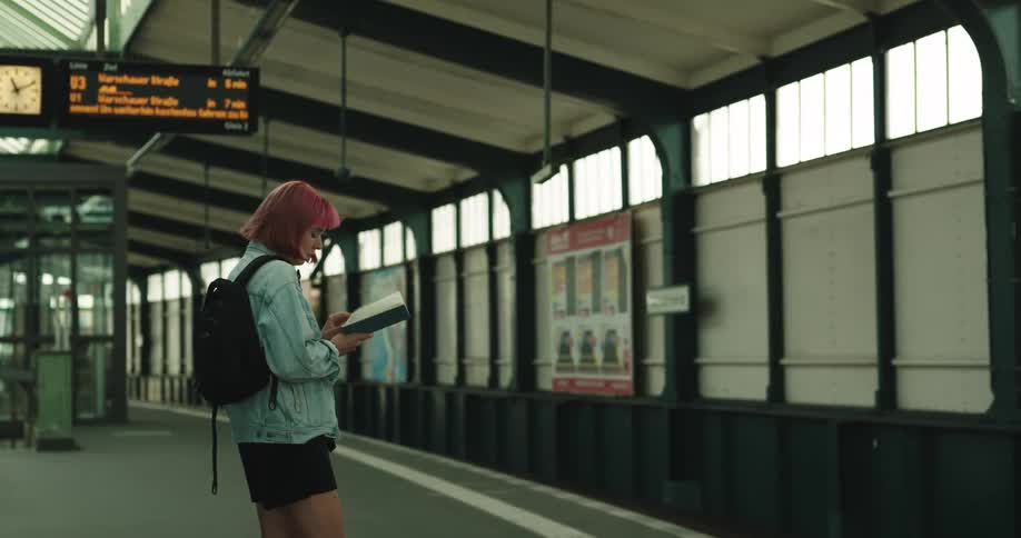 A girl with wine colored hair and wearing a backpack is standing in a train station reading a book