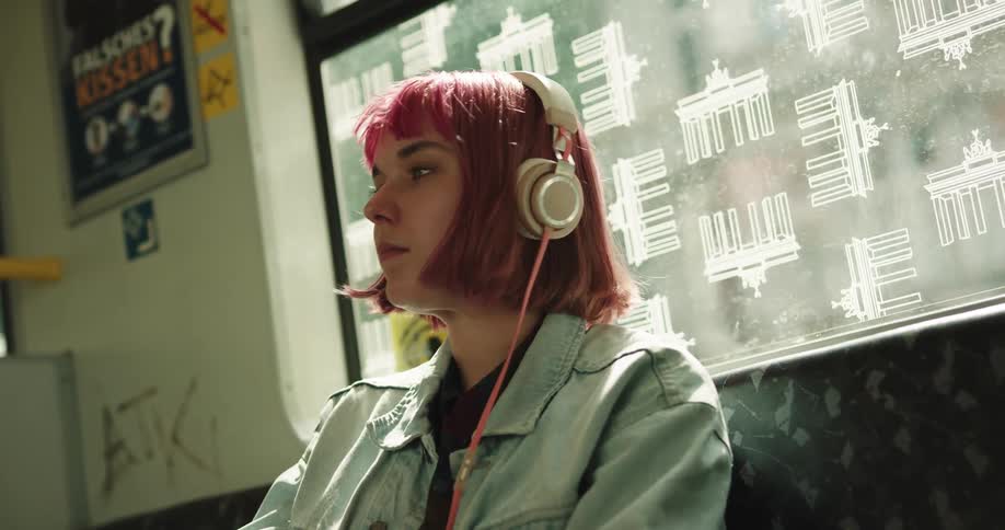 A girl with wine colored hair and headphones on her head is sitting on a train listening to music