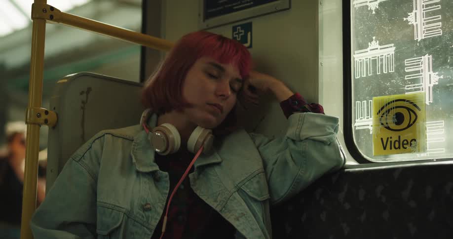 Woman with dyed hair wearing headphones sits near open door of public transit car as passenger walk by
