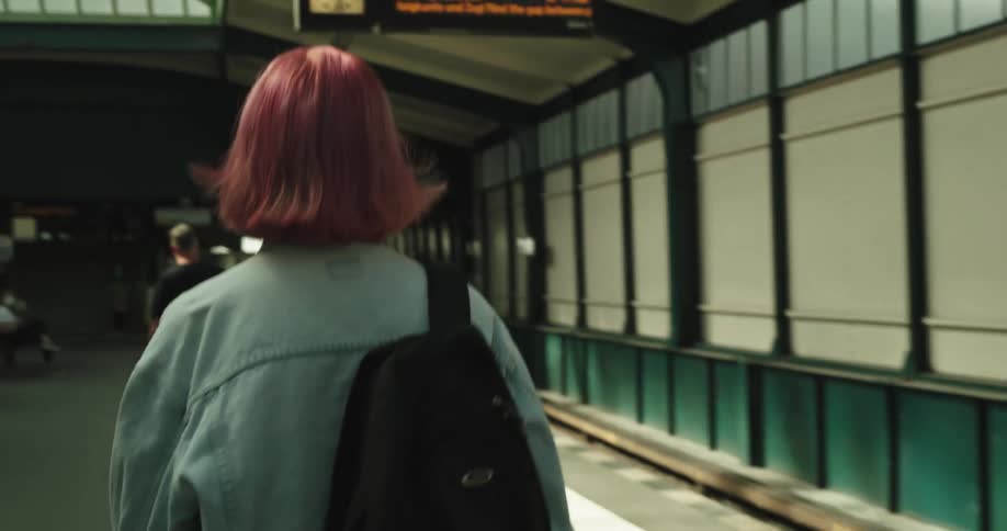 Woman with dyed hair carrying backpack walks along transit terminal towardtracks