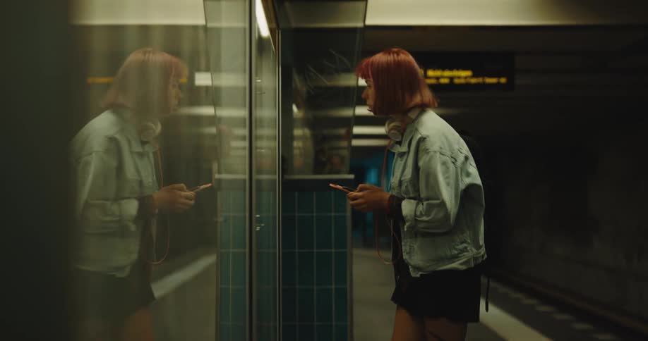 A woman with headphones and a smart phone in a train station with a sign in the background
