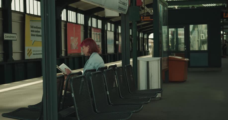 A woman sitting on a chair reading a book on a train station with signs in the background