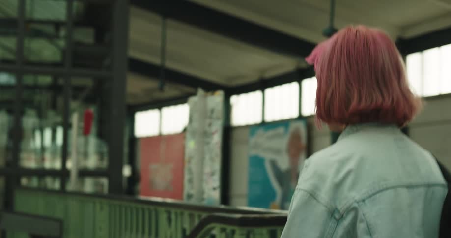 Woman with pink hair denim jacket black backpack and shorts walks in train station