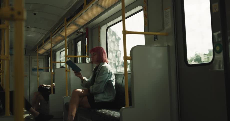 A woman is sitting on a train as it moves and is reading a book