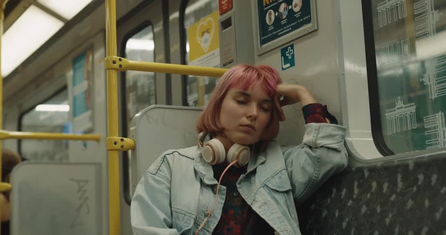 A girl with wine colored hair and headphones around her neck is sitting on a train leaning her head on her hand