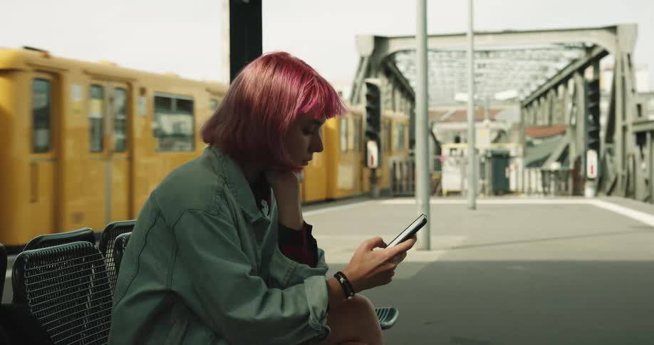 Woman with dyed hair sits at transit scrolling on mobile phone as yellow train passes