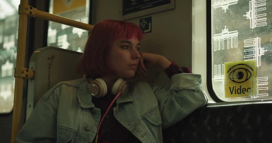 Woman with hot pink hair wearing headphones sits on public transport looking out window as nearby doors close and train leaves the station