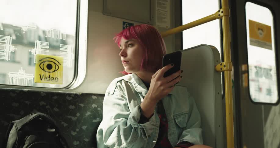 A woman sitting on a train next to a backpack looking at a smart phone