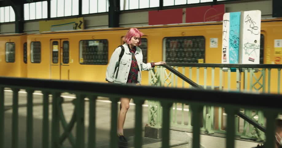 A yellow trains stops and a girl with wine colored hair and a backpack gets off and walks down stairs