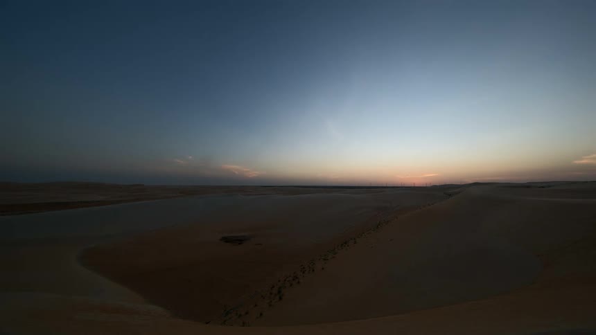 A breeze blows sand off dunes in the desert during sunset