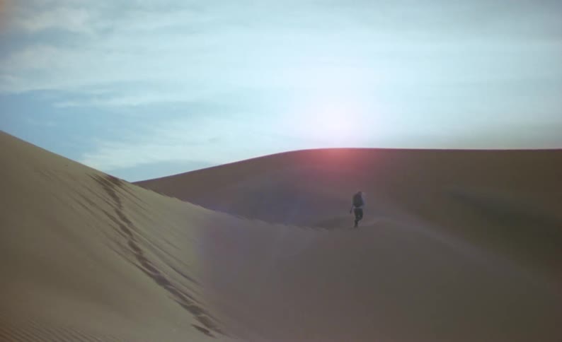 A person with a backpack walking in a desert with the sky and clouds in the background