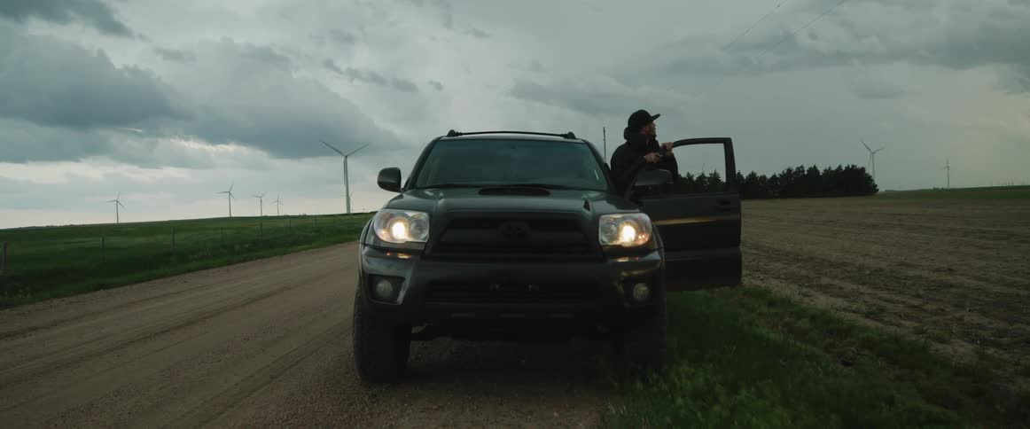 Man holds onto open door of vehicle parked on side of dirt road as he looks out across field with wind farm in background