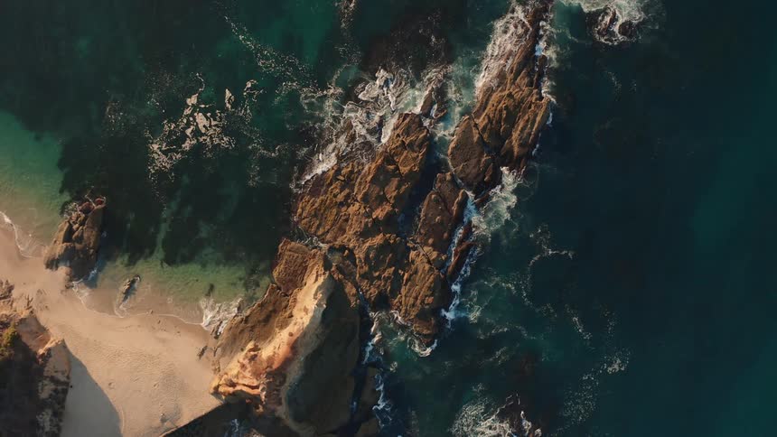 Aerial view of a shoreline with waves rolling over a beach and splashing against rocks