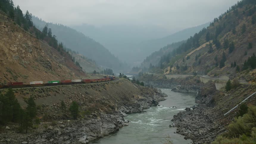 A river next to a train trees and cliffs with the sky and clouds in the background