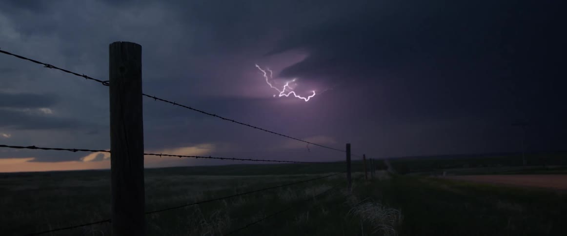 Lighting strikes through storm clouds on horizon in background with barbed wire and posts border dirt road in foreground