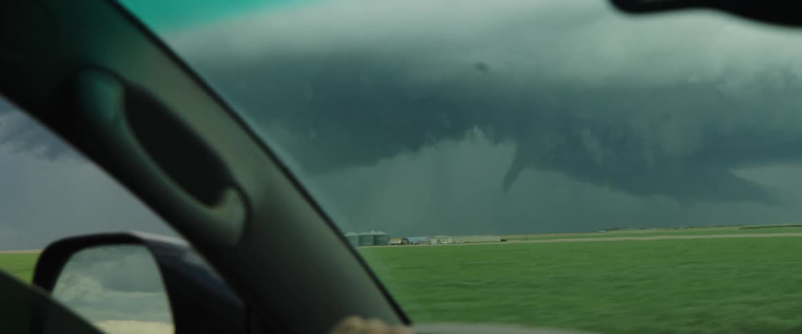 A vehicle travels on a road next to a grassland with thunder clouds looming overhead